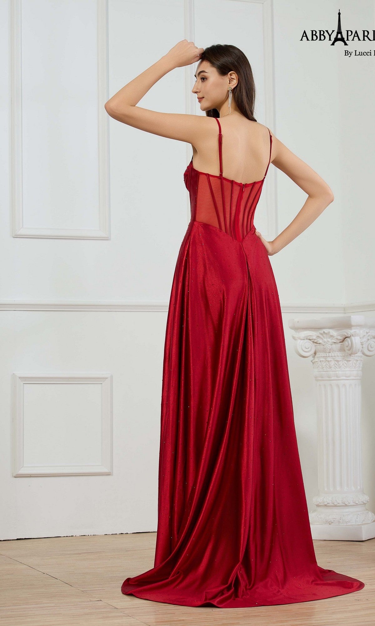A woman wearing the Abby Paris 90374 long prom dress, featuring metallic stretch satin and a sheer back panel, stands indoors facing away from the camera.
